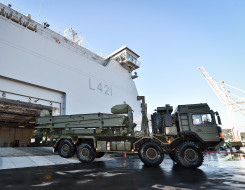 Royal New Zealand Navy's HMNSZ Canterbury berthed as a New Zealand Army MHOV drives off the ship. In the background there is blue sky and a crane as part of the port in Wellington. 