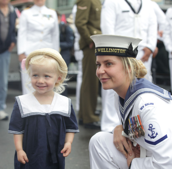 A sailor bends down to engage with a young child who is dressed in a sailor outfit. They are both smiling. You can also see other sailors standing in the background of the photo