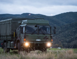 A New Zealand Army Medium and Heavy Operational Vehicle (MHOV) with lights on sits in a grassy area with a green covered hill in the background. It's a darker image with grey sky.