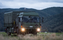 A New Zealand Army Medium and Heavy Operational Vehicle (MHOV) with lights on sits in a grassy area with a green covered hill in the background. It's a darker image with grey sky.