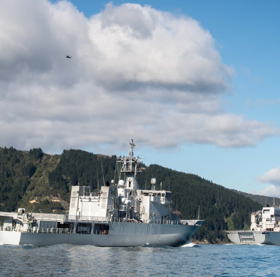 HMNZS Wellington transits behind HMNZS Canterbury in the Marlborough Sounds on a nice day - clouds and blue sky. Green hill area in the background the photo. 