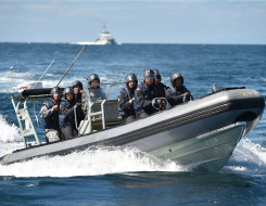 A Royal New Zealand Navy Rigid-Hulled Inflatable Boat (RHIB) full with sailors wearing helmets moves through the ocean with a ship in the background on a nice day, blue sky.