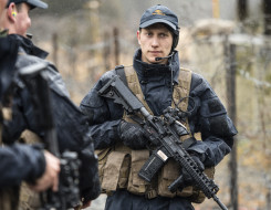 A Royal New Zealand Air Force airman carrying a MARS-L weapon wearing a headset and Air Force cap. To the left in the foreground two other airmen look away from the camera and are out of focus. They are wet from the rain. In the background you can see a b