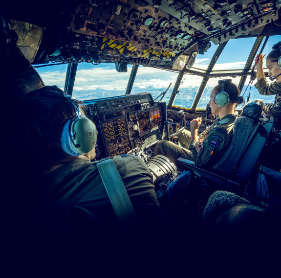 The Royal New Zealand Air Force aircrew on board one of the C-130H(NZ) Hercules aircraft during a flight. Outside the window of the aircraft you can see land and a sunny day with some cloud. Within the flight deck there is a pilot close to the left side o