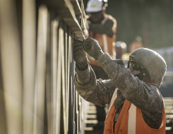 A New Zealand Army soldier works on a bridge with a helmet, glasses and an orange high vis vest. There are other people in the background working on the bridge as well.