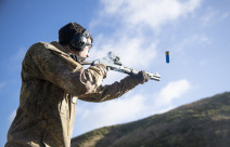 A soldier fires a Benelli M3 (NZ) Tactical Shotgun. The photo is shot from a low angle looking up to the soldier from the side. The soldier is wearing hearing protection and you can see the bullet coming out. There is a hill in the background. 