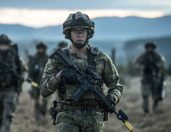 A New Zealand Army soldier in uniform in the foreground carries a MARS-L weapon with fellow soldiers in the background