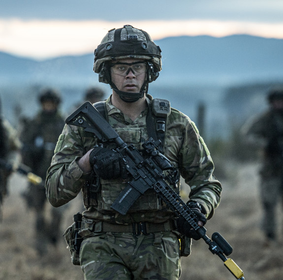 A New Zealand Army soldier in uniform in the foreground carries a MARS-L weapon with fellow soldiers in the background