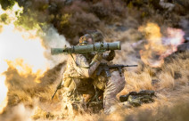 New Zealand Army soldiers fire a Carl Gustaf M3 there are flames out the back and motion shown out the front of the weapon. the soldiers are training in the tussock grass