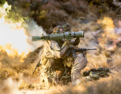 New Zealand Army soldiers fire a Carl Gustaf M3 there are flames out the back and motion shown out the front of the weapon. the soldiers are training in the tussock grass