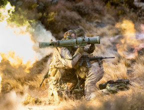 New Zealand Army soldiers fire a Carl Gustaf M3 there are flames out the back and motion shown out the front of the weapon. the soldiers are training in the tussock grass