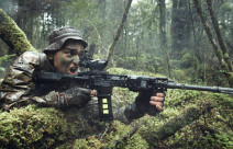 A New Zealand Army soldier in the New Zealand bush with MARS-L weapon. The area is all wet and very green. The soldier has his mouth open as he yells a command. 