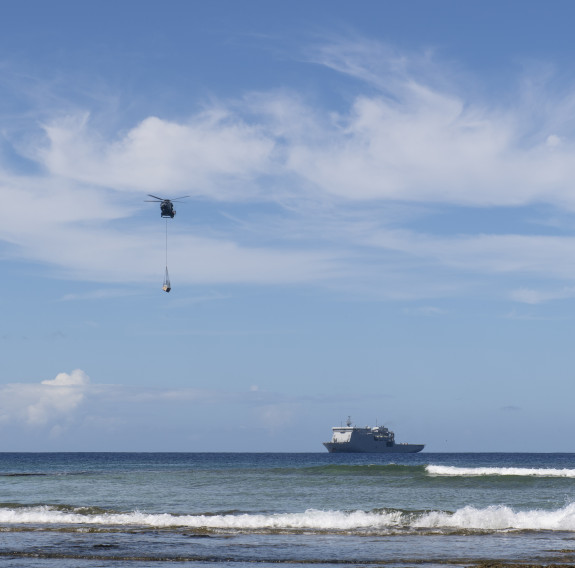 A Royal New Zealand Air Force NH90 helicopter carries an underslung load near HMNZS Canterbury out in the ocean on a nice day. 