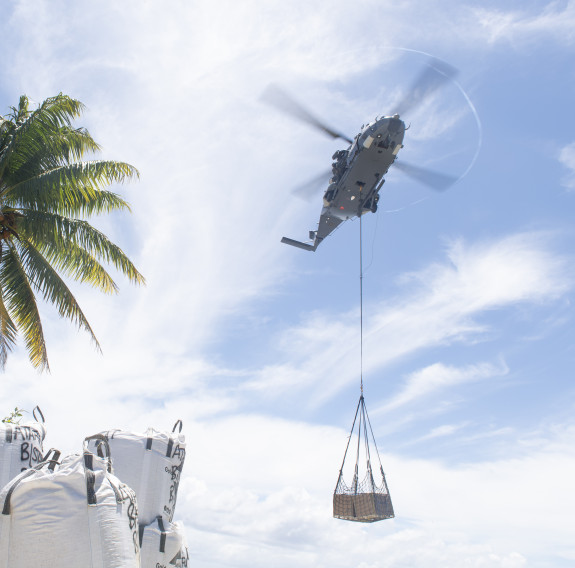 A Royal New Zealand Air Force NH90 helicopter carries an underslung load on the right of the frame. On the left of the frame there are palm trees and other loads.