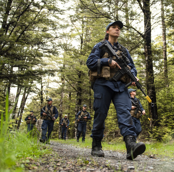 A Royal New Zealand Air Force airman walks with a weapon on a shingled path with other airmen in the background. 