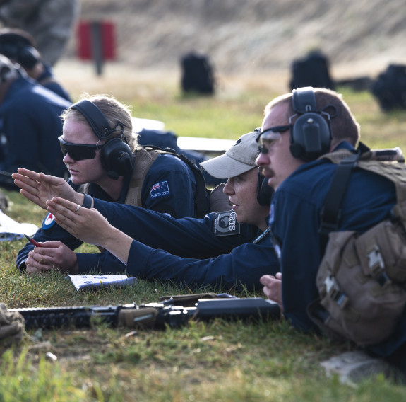 Royal New Zealand Air Force personnel work together on the rifle range. One of the personnel signals in the direction to the left while the other two look in that direction. All personnel are wearing hearing protection.