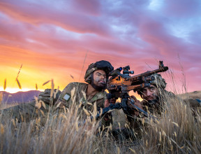 Two New Zealand Army soldiers lie in the tussock grass with the MAG 58 - 7.62mm Machine Gun. One soldier looks through the scope and the other looks toward the target. The sky is pink and yellow sunset skies. 