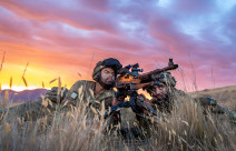 Two New Zealand Army soldiers lie in the tussock grass with the MAG 58 - 7.62mm Machine Gun. One soldier looks through the scope and the other looks toward the target. The sky is pink and yellow sunset skies. 