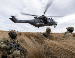 New Zealand Army soldiers sit in the tussock grass within the Waiouru Military Training Area after being dropped off by an NH90 helicopter (seen behind them flying away). 