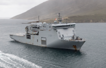 HMNZS Canterbury sailing in the ocean, a Rigid Hull Inflatable Boat (RHIB) moves toward the ship from the bottom of the image. The day looks dreary and cloudy.