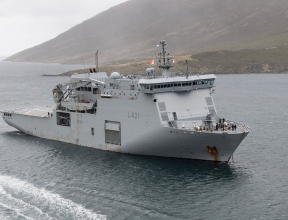 HMNZS Canterbury sailing in the ocean, a Rigid Hull Inflatable Boat (RHIB) moves toward the ship from the bottom of the image. The day looks dreary and cloudy.