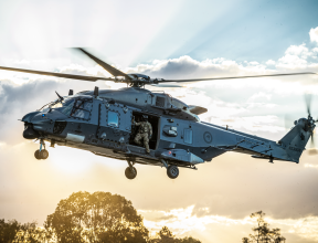 An NH90 helicopter flies in the sky with the helicopter loadmaster looking out the door to check the ground before landing. The sun is streaming behind the helicopter and cloud behind it. 