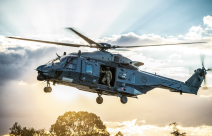 An NH90 helicopter flies in the sky with the helicopter loadmaster looking out the door to check the ground before landing. The sun is streaming behind the helicopter and cloud behind it. 