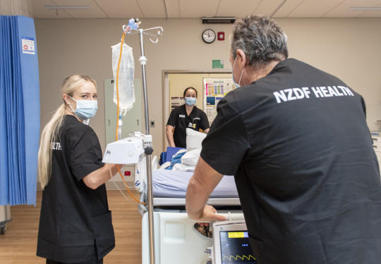 Three nurses move a medical bed in a hospital environment. They are all wearing black scrubs that read 'NZDF HEALTH' on the back. In the background is a doorway to a hall and there is a blue curtain hanging from the ceiling on the left.