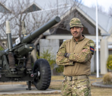 Staff Sergeant TJ Chapman, wearing fatigues standing cross armed in front of an L119, 105mm Light Gun, which is parked outside a headquarters building.