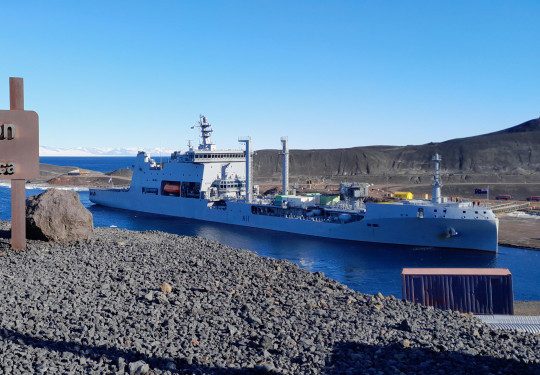 HMNZS Aotearoa arriving at McMurdo Station in Antartica behind a metal sign that reads 'McMurdo Station Antartica.'