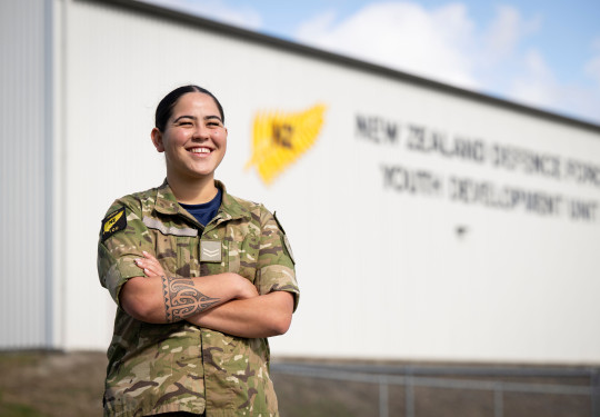 A woman with a traditional tattoo on her forearm stands with arms crossed while wearing military uniform. She smiles as she looks into the distance in front of a large grey building with words that are blurred out on the side.
