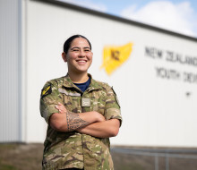 A woman with a traditional tattoo on her forearm stands with arms crossed while wearing military uniform. She smiles as she looks into the distance in front of a large grey building with words that are blurred out on the side.