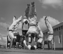 A black and white photo of Physical Training Instructors showcase their strength and agility in a gymnastic display in the 1960s. They conduct handstands on the ground and various aparatus.