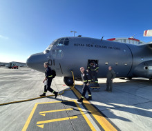 People disembark a Hercules aircraft, walking out the door to the front of the aircraft. Blue sky in the background. 