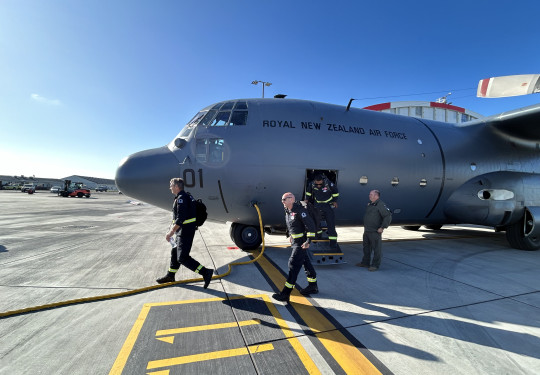 People disembark a Hercules aircraft, walking out the door to the front of the aircraft. Blue sky in the background. 