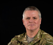 Army soldier stands in uniform, crossed arms, staring at the camera. 