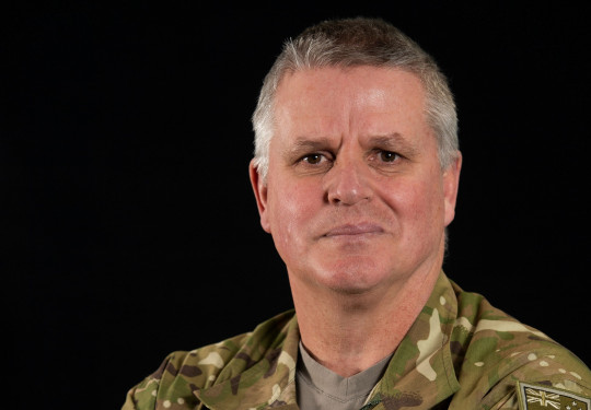 Army soldier stands in uniform, crossed arms, staring at the camera. 