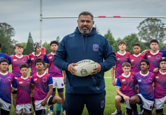 A man holds a rugby ball in front of a team of young players.