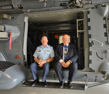 Two people sit in the side door of an NH90 helicopter in a hangar.