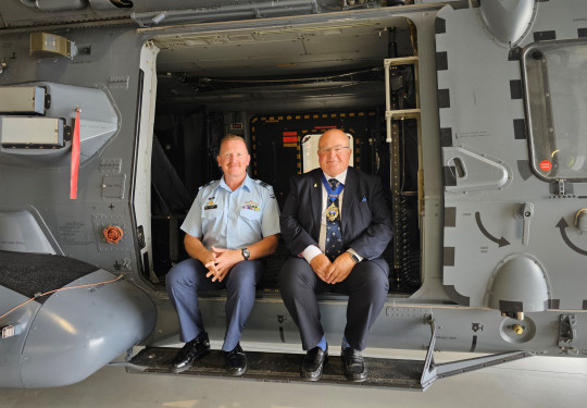 Two people sit in the side door of an NH90 helicopter in a hangar.