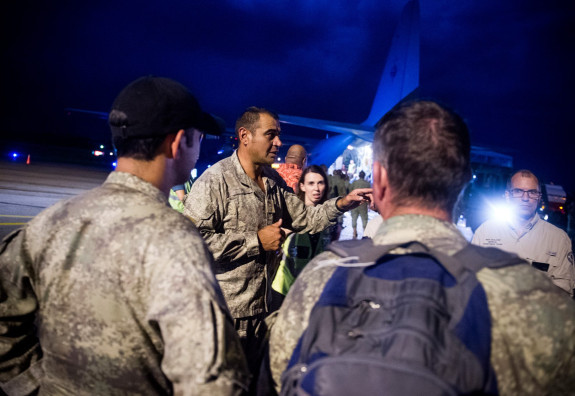 Three New Zealand soldiers in dress fatigues and a man from another government agency are the visible part of a huddle on the tarmac of an airport at night, two soldiers have their back to the camera, they frame te third soldier who is speaking and gestur