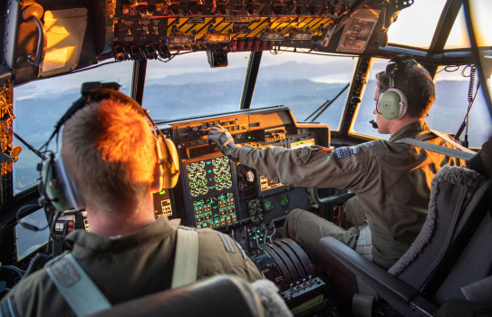 View from the Hercules aircraft flight deck as the crew transport military aid between staging centres in Europe. In the image there are two RNZAF personnel (pilot and co-pilot). There is sunlight in the flight deck. 