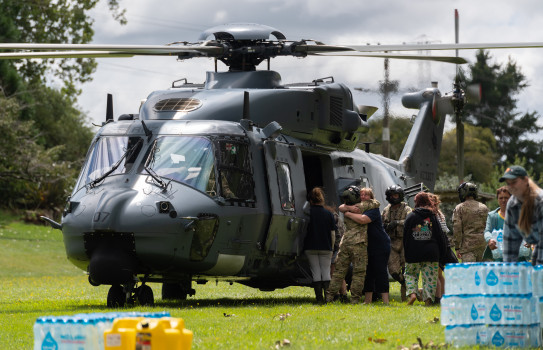 Supplied being unloaded from a NH90 helicopter in a field with trees in the background and bottled water and supplies in the foreground.