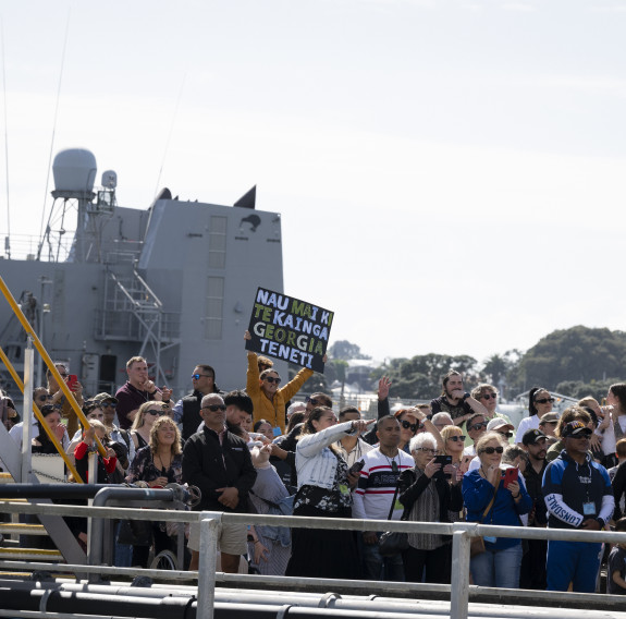 Family and friends wave and hold signs on a wharf. Part of another Navy ship is visible in the background.