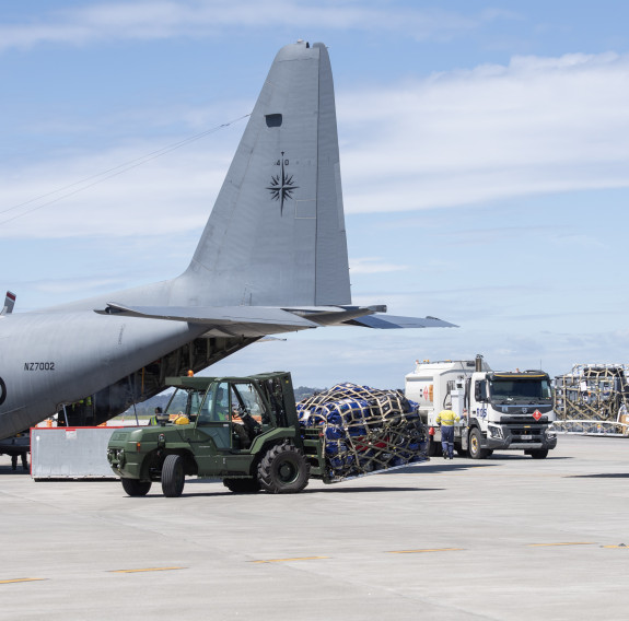 A military green forklift carries a pallet of aid, secured with webbing on the tarmac of an airfield. The tail end end of a C-130H Hercules with cargo ramp open is visible in the background.