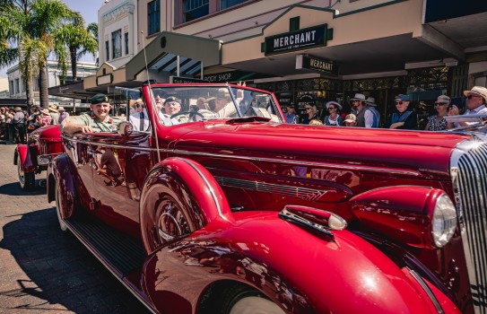 A large red vehicle drives down a street flanked with members of the public dressed in art deco era clothing, the vehicle holds army staff smiling and waving at the crowds.