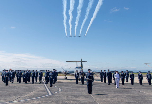 Royal New Zealand Air Force personnel standing in a formation on the flight line on a nice blue sky day. Five T-6C Texan II aircraft fly overhead and a King Air 350 aircraft sits in the background.