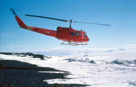 An orange Iroquois helicopter takes off over the mountainous terrain of Antarctica under blue skies.