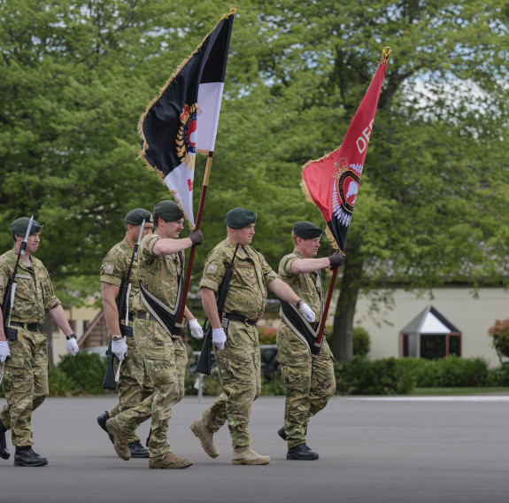 Five soldiers march in front of a building and trees, two bearing flags.