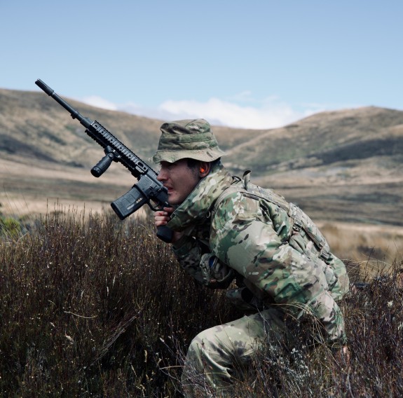 A soldier holds a weapon as they move through tussocks in front of rolling hills.